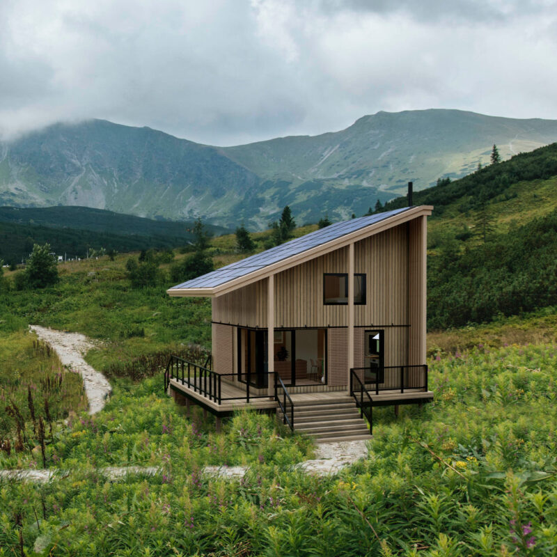 A modern wooden CABN with a sloped roof and solar panels sits along a winding stone path in a lush green mountain valley, with dramatic peaks and low-hanging clouds in the background.