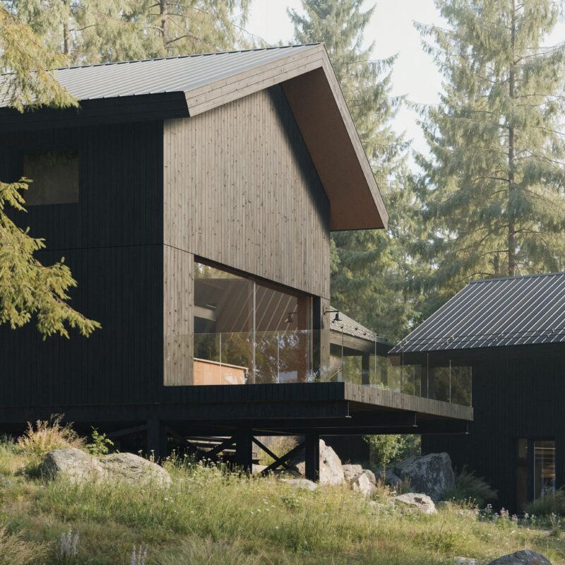 A modern black wooden cabin with a sloped metal roof and large glass windows, elevated on stilts with a glass-railing deck, set in a forested area surrounded by tall pine trees, rocks, and wild grasses. Design in collaboration with Altruist Built and CABN.